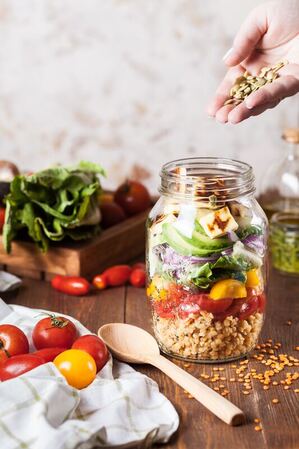 Person holding brown grains over a jar full of fresh salad makings.