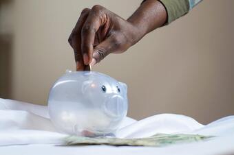 A hand putting coins in a mostly empty translucent piggy bank.