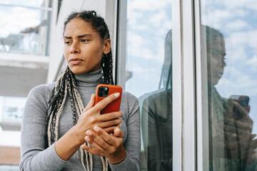 Woman leaning against a window with a perplexed look on her face, while holding a smart phone.