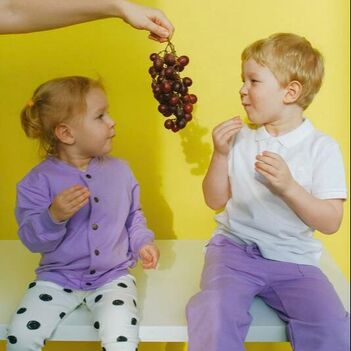 Girl and boy sitting on white table eating grapes.